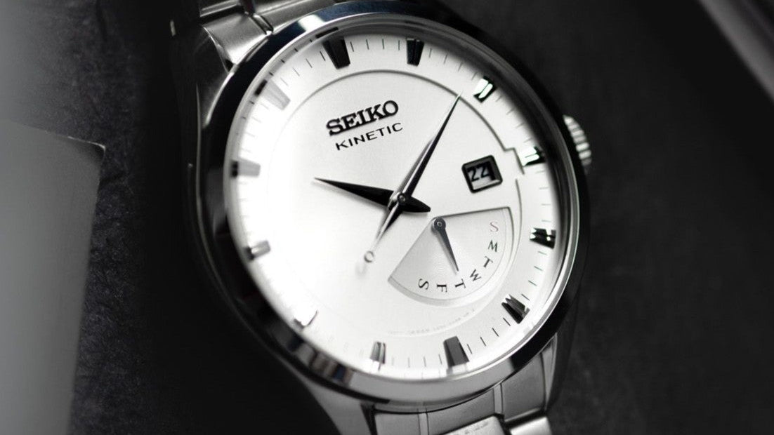 A close-up shot of a Seiko Kinetic watch with a silver stainless steel case and bracelet, featuring a white dial with black markings and hands.