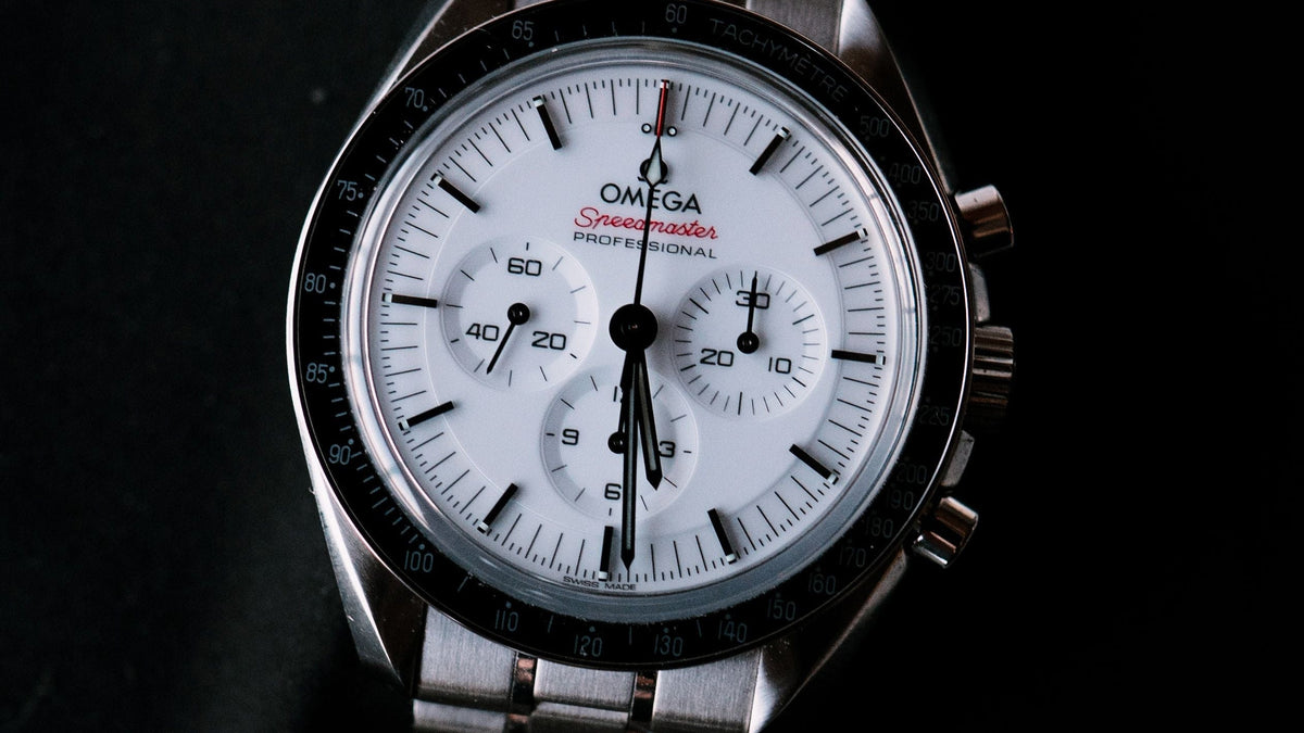 Omega x Swatch Speedmaster Cosmograph Daytona wristwatch with a silver stainless steel bracelet, black bezel, and white dial on a dark background.