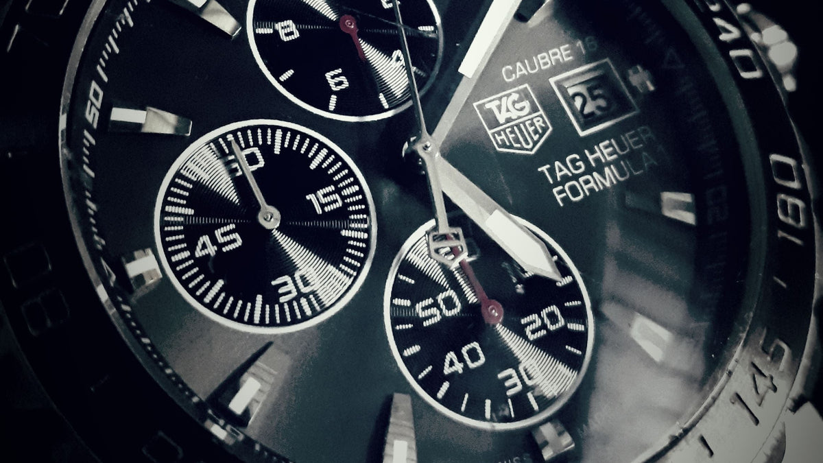 A close-up shot of a Tag Heuer Calibre chronograph watch with a black dial, silver-tone bezel, and a matching stainless steel link bracelet, set against a dark background.